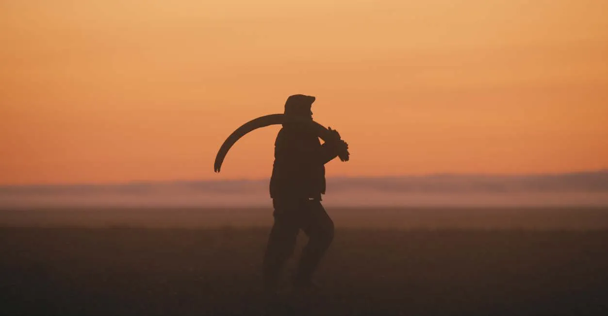 A figure carrying a mammoth tusk in silhouette against an orange sunset.